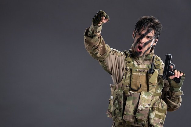 Vista frontal del joven soldado de camuflaje con pistola en la pared oscura