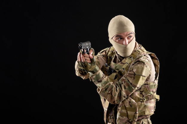 Vista frontal del joven soldado en uniforme apuntando con pistola en la pared negra