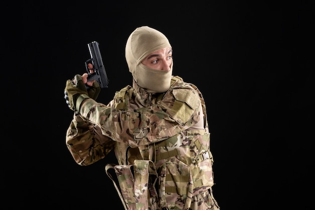 Vista frontal joven soldado en uniforme con pistola en pared negra