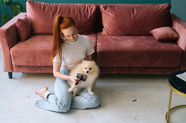 Vista frontal de una joven sonriente peinando suavemente a un pequeño perro mascota Spitz bastante blanco sentado en el suelo en casa