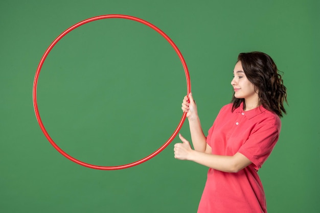 Vista frontal joven vendedora sosteniendo hula hoop rojo sobre fondo verde cuerpo de yoga uniforme trabajo trabajo compras venta