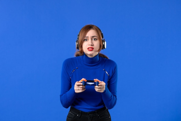 Vista frontal de la jugadora con auriculares y gamepad jugando videojuegos sobre fondo azul niña sofá juvenil reproductor de video para adultos ganador de alegría
