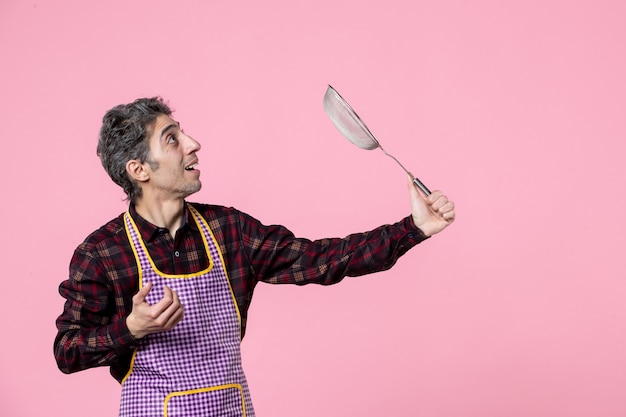 Vista frontal macho joven en el cabo sosteniendo el tamiz y preparándose para golpear algo sobre fondo rosa profesión color cocinero marido trabajador horizontal comida uniforme de cocina