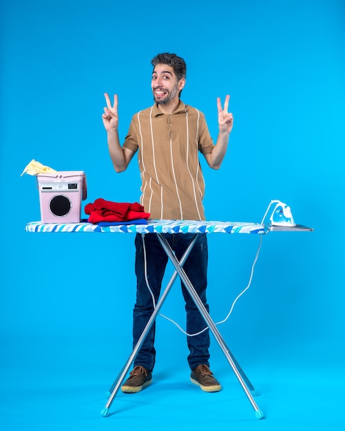 Vista frontal macho joven detrás de la tabla de planchar posando y sonriendo sobre fondo azul color de las tareas del hogar lavandería hombre emoción limpia lavadora