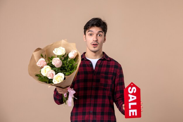 Vista frontal macho joven sosteniendo hermosas flores para mujer y placa de venta en pared marrón