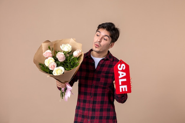 Foto vista frontal macho joven sosteniendo hermosas flores y placa de venta en pared marrón