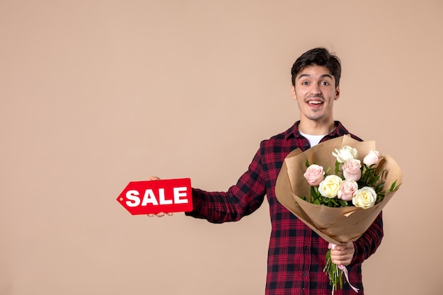 Vista frontal macho joven sosteniendo hermosas flores y placa de venta en pared marrón