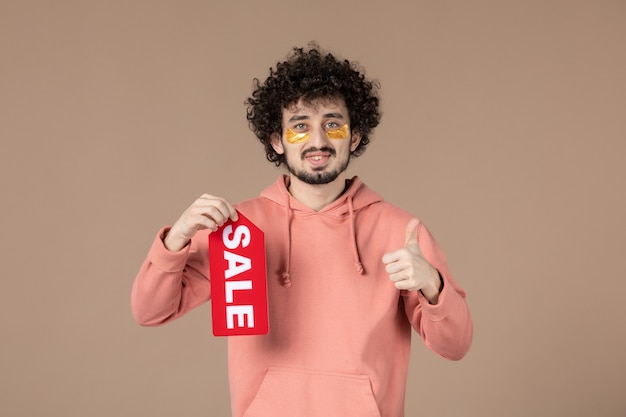 Vista frontal macho joven sosteniendo placa de venta sobre fondo marrón salón de compras de piel terapia de cuidado de la piel masaje facial