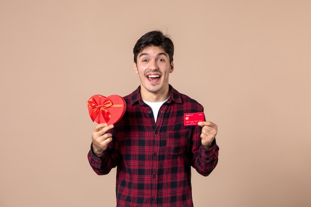 Vista frontal macho joven sosteniendo presente en forma de corazón y tarjeta bancaria en la pared marrón