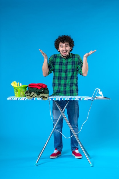Vista frontal macho joven con tabla de planchar sobre fondo azul quehaceres domésticos plancha ropa limpia casa lavadora