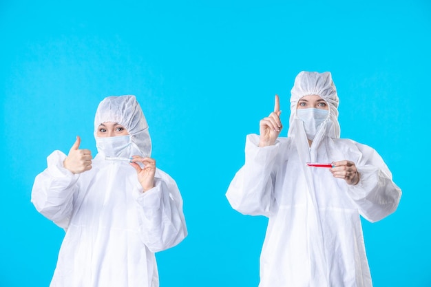 vista frontal médicas femininas em trajes protetores e máscara segurando frascos sobre o fundo azul covid- saúde ciência hospital vírus pandêmico cor