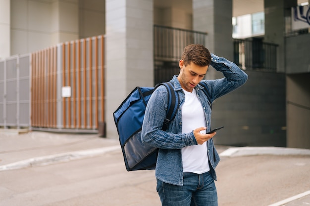 Vista frontal del mensajero masculino guapo confundido con mochila termo usando la aplicación de navegación en el teléfono de pie en la calle de la ciudad. Repartidor en busca de la dirección del cliente en busca de smartphone.