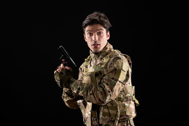 Vista frontal del militar en uniforme sosteniendo la pistola en la pared negra