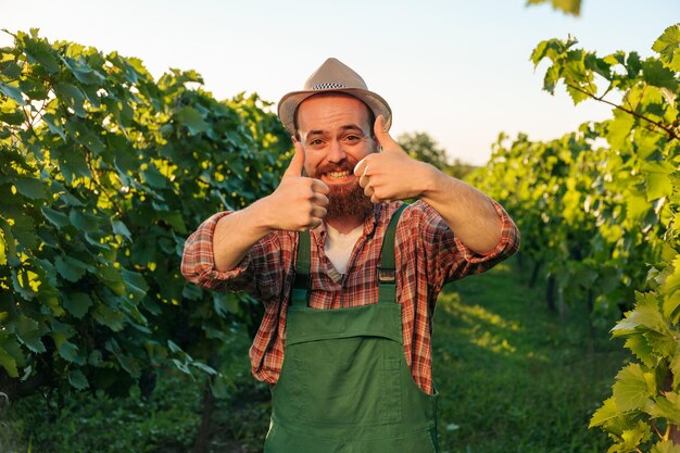 Vista frontal mirando a la cámara joven enólogo agricultor trabajador feliz sonrisa ampliamente ambas manos pulgar arriba