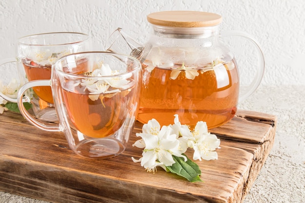 Vista frontal de una moderna taza de vidrio de doble fondo con una deliciosa tetera de vidrio de té de jazmín y flores en una tabla de madera