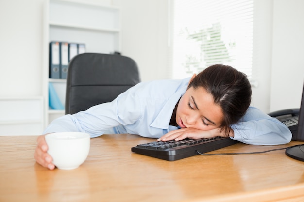 Vista frontal de una mujer bonita durmiendo en un teclado mientras sostiene una taza de café