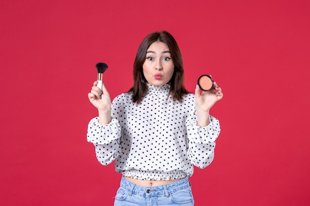 Vista frontal de la mujer joven con borla y polvo haciendo maquillaje en la pared roja