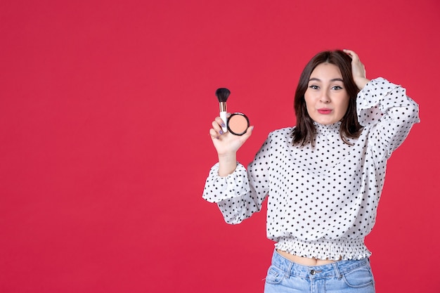 Vista frontal mujer joven con borla en polvo para maquillaje y pared roja