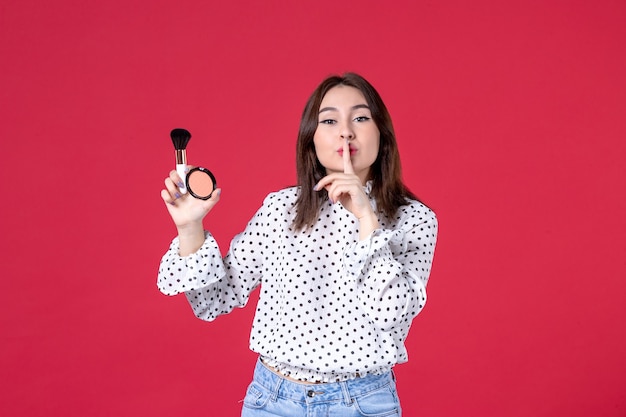 Vista frontal de la mujer joven con borla y polvo para maquillaje en pared roja