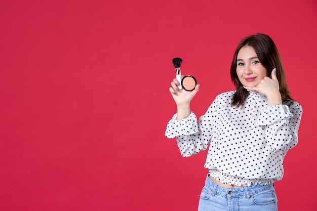 Vista frontal de la mujer joven con borla y polvo para maquillaje en pared roja