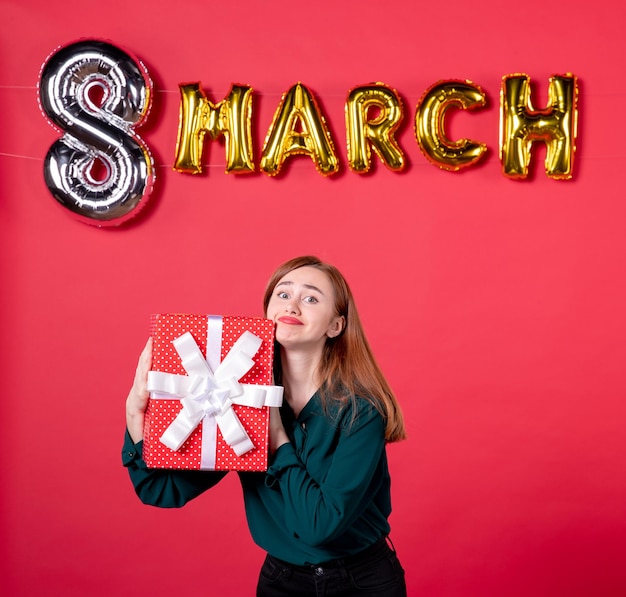 Vista frontal mujer joven con decoración de marzo y presente sobre fondo rojo vacaciones de glamour femenino sensual día de la mujer moda mujer regalo