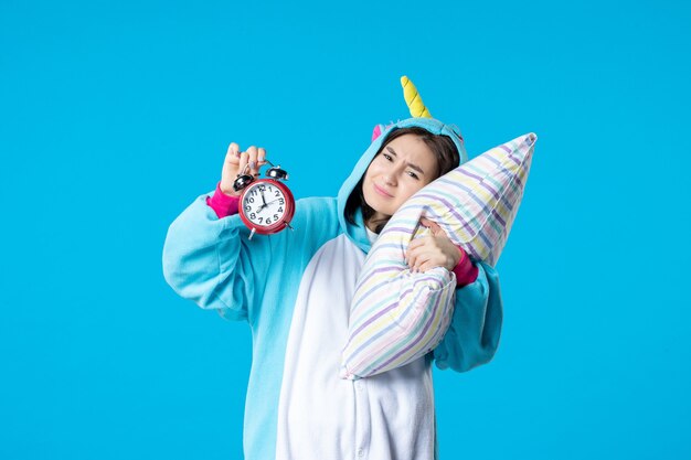 Vista frontal mujer joven en fiesta de pijamas sosteniendo almohada y relojes sobre fondo azul sueño dormir hasta tarde descanso pesadilla cama noche diversión bostezo