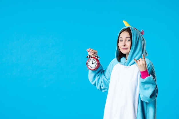 Vista frontal mujer joven en fiesta de pijamas sosteniendo relojes rojos sobre fondo azul dormir descanso pesadilla divertido juego mujer cama sueño tarde hora