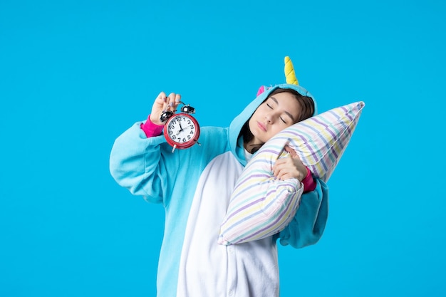 Vista frontal mujer joven en fiesta de pijamas sosteniendo relojes y tratando de conciliar el sueño en la almohada sobre fondo azul cama sueño dormir hasta tarde descanso pesadilla noche amigos divertidos bostezo