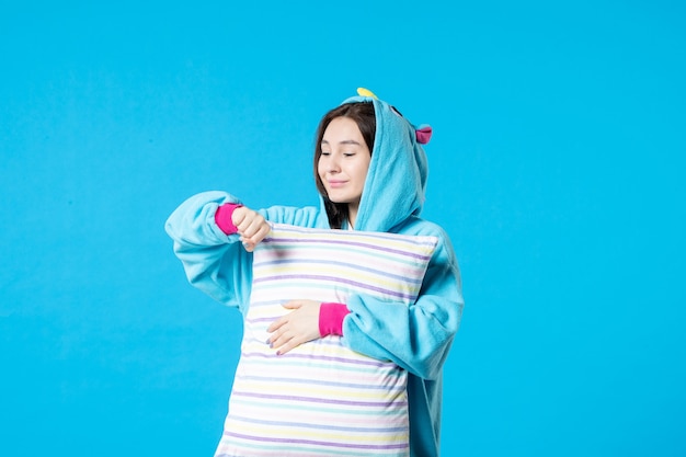 Vista frontal mujer joven en pijama para fiesta de pijamas sosteniendo almohada sobre fondo azul cama mujer descanso nocturno pesadilla tarde diversión sueño