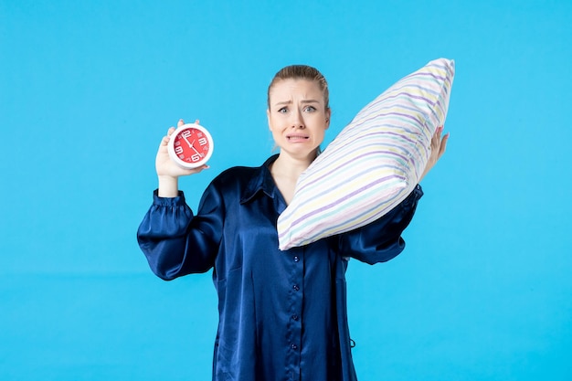 Foto vista frontal mujer joven en pijama con reloj y almohada sobre fondo azul noche cansada fiesta descanso sueño sueño cama pesadilla tarde