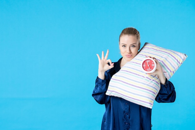 Vista frontal mujer joven en pijama sosteniendo relojes y almohada sobre fondo azul cama cansada descanso nocturno dormir tarde mujer fiesta sueño