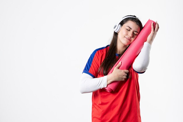 Vista frontal mujer joven en ropa deportiva con estera de yoga escuchando música pared blanca