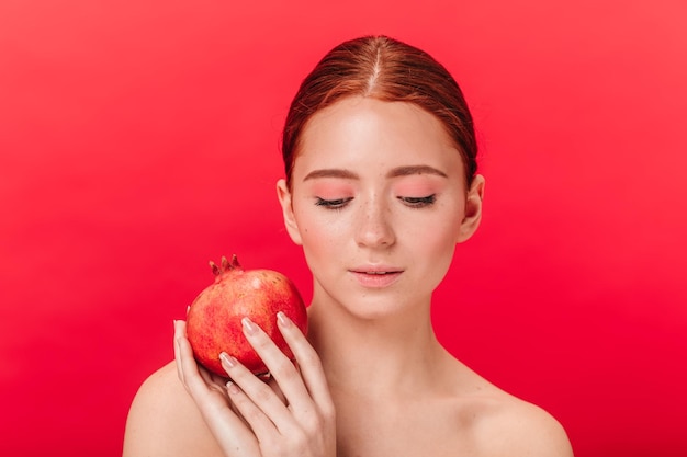 Vista frontal de la mujer pelirroja sosteniendo granate con los ojos cerrados Foto de estudio de la modelo femenina desnuda con granada