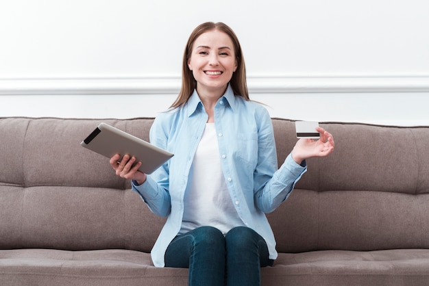 Foto vista frontal mujer sentada en el sofá con tableta digital y tarjeta de crédito