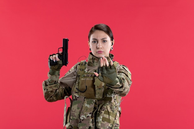 Vista frontal de la mujer soldado en camuflaje con pistola en la pared roja