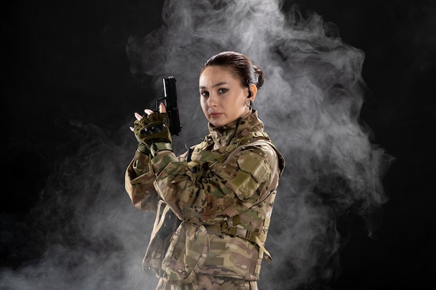 Vista frontal de la mujer soldado en uniforme sosteniendo la pistola en la pared negra