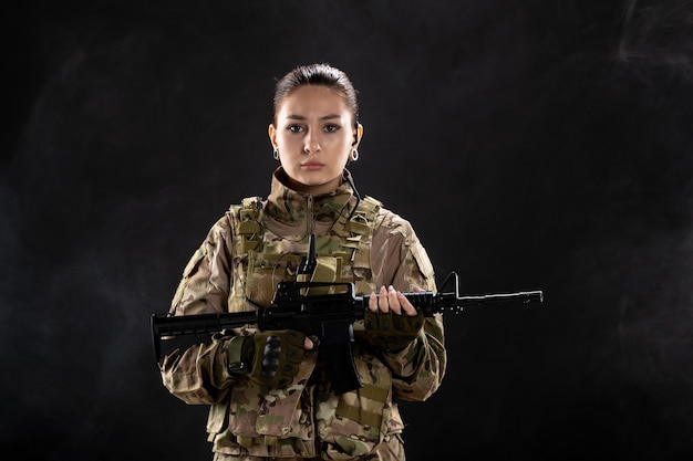 Vista frontal mujer soldado en uniforme sosteniendo rifle en pared negra