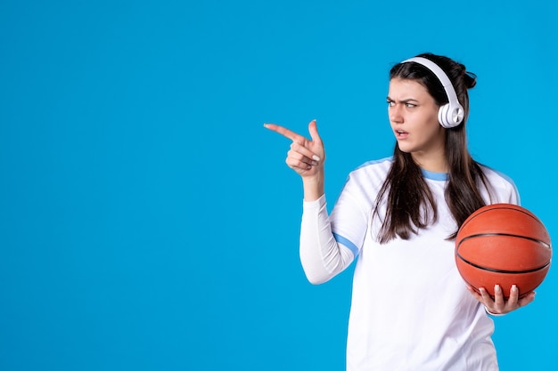 Vista frontal de las mujeres jóvenes con auriculares sosteniendo baloncesto en la pared azul