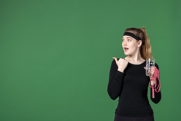 Vista frontal de las mujeres jóvenes sosteniendo la medida de la cintura y un vaso de agua sobre fondo verde, color, salud, cuerpo, entrenamiento, atleta, deporte, negro