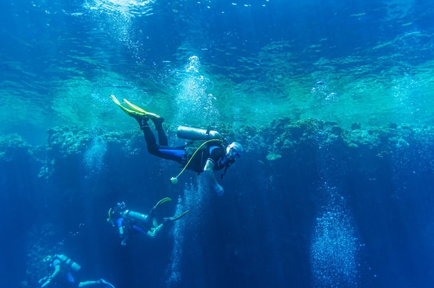 Vista frontal de la natación del grupo de buzos que exploran el agua azul del océano profundo y oscuro con el telón de fondo de un arrecife de coral. Macho y hembra en aletas examina el fondo del mar. Bucear. Vida activa Burbujas de aire.