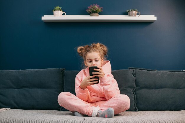 Vista frontal de una niña rubia sentada en casa en el sofá con un teléfono móvil para niños y aparatos