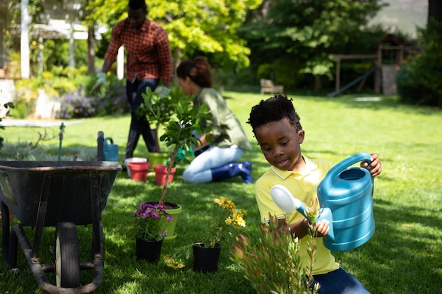 Vista frontal de un niño afroamericano en las plantas de riego del jardín, con sus padres haciendo jardinería en el fondo. Familia disfrutando del tiempo en casa, concepto de estilo de vida