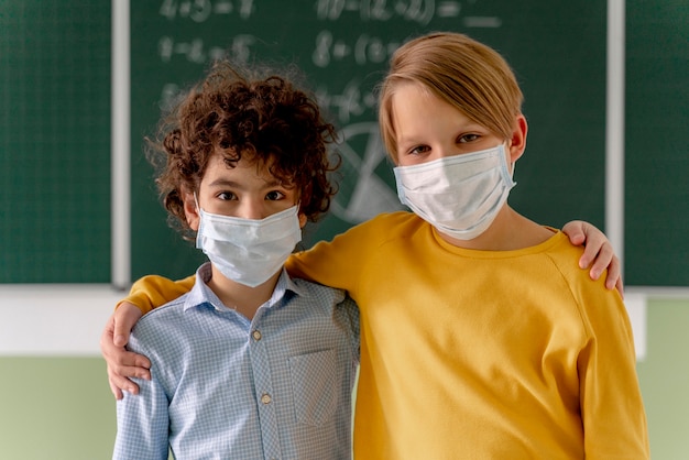 Vista frontal de los niños con máscaras médicas posando en el aula frente a la pizarra