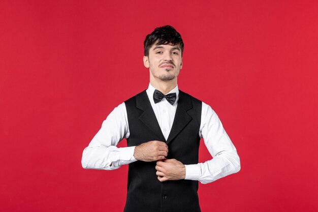 Foto vista frontal del orgulloso camarero masculino en uniforme con mariposa en el cuello en la pared roja