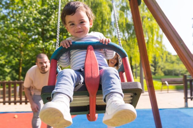 Foto vista frontal de padres balanceando a un niño pequeño en el patio de recreo en un día soleado