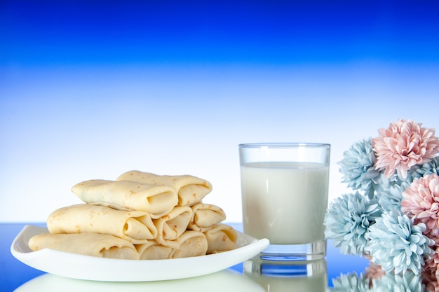 Vista frontal de panqueques dulces enrollados con un vaso de leche sobre fondo azul claro pastel de comida desayuno color comida mañana flor de azúcar