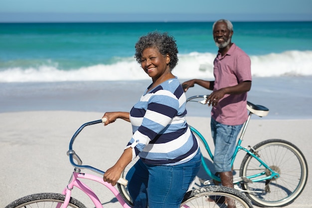 Vista frontal de una pareja afroamericana de alto nivel parada en la playa con el cielo azul y el mar al fondo, conduciendo sus bicicletas y sonriendo a la cámara