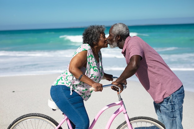 Vista frontal de una pareja de ancianos afroamericanos besándose en la playa, la mujer en una bicicleta, con el cielo azul y el mar en el fondo