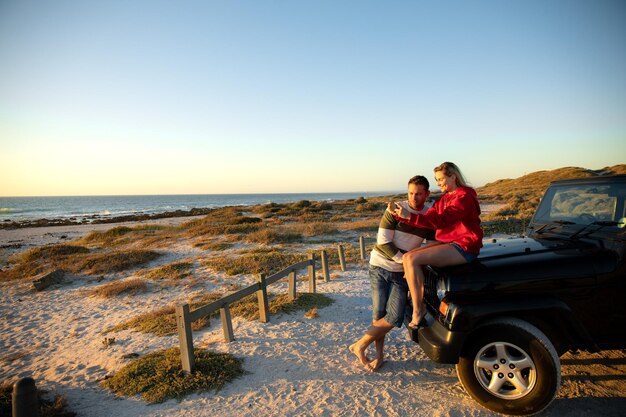 Vista frontal de una pareja caucásica afuera de su auto abierto, abrazándose y mirando el mar. Vacaciones de fin de semana en la playa, estilo de vida y ocio.