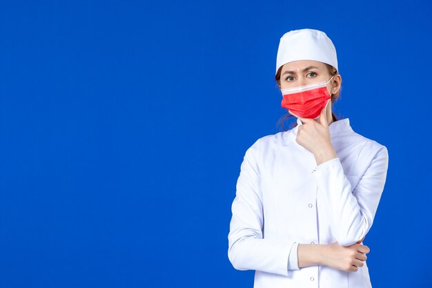 Vista frontal pensando joven enfermera en traje médico con máscara roja sobre una pared azul